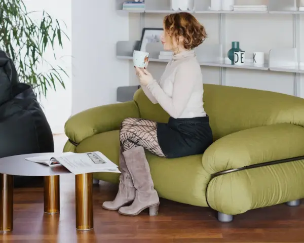 Donna sorseggia una bevanda su un divano verde, circondata da arredi moderni. Un pouf nero e un tavolino completano l'ambiente, creando un'atmosfera accogliente con divani in stile.