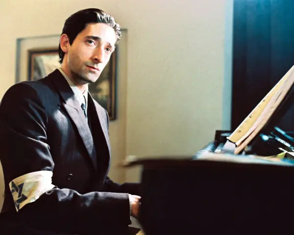 Adrien Brody jako Władysław Szpilman w filmie "Pianista" Romana Polańskiego, z opaską na ramieniu, siedzi przy fortepianie.