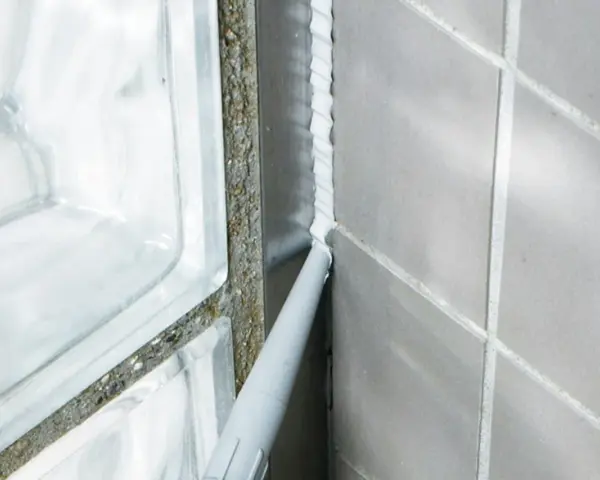 Applicator sprutar ny silikonfog längs en fog i badrummet, bredvid en duschvägg av glasblock.