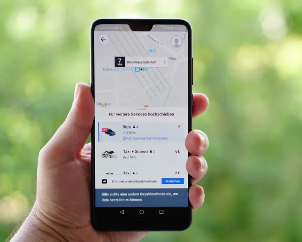 Hand hält Smartphone mit der besten Taxi App Deutschlands. Anzeige zeigt Fahrtoptionen nach Köln Hauptbahnhof.