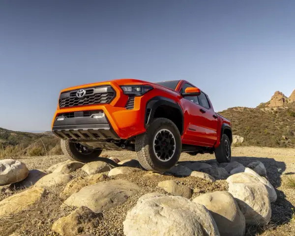 Pomarańczowy Toyota pickup pokonuje kamienisty teren na tle wzgórz.