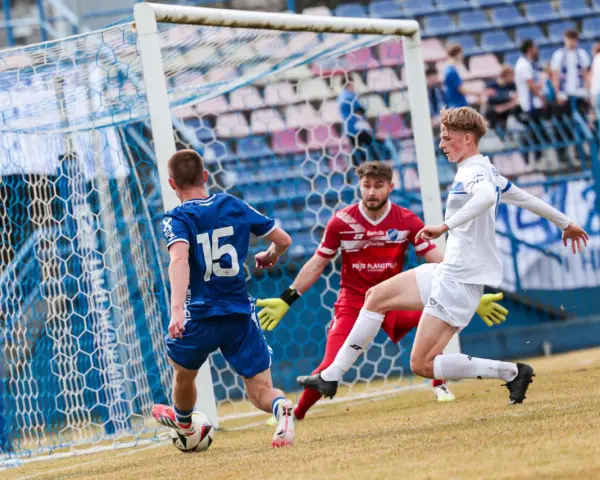 Piłkarz Michał Bartczak w białej koszulce próbuje strzelić gola, obrońca w niebieskiej koszulce z numerem 15 i bramkarz w czerwonej próbuje go zatrzymać.