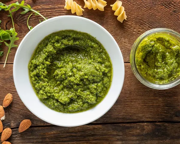 Pesto di rucola e mandorle in una ciotola bianca e un vasetto, con mandorle e rucola fresca intorno.