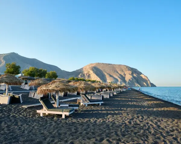 Czarne piaski i leżaki pod słomianymi parasolami na Santorini plażach. W tle góry i spokojne morze.