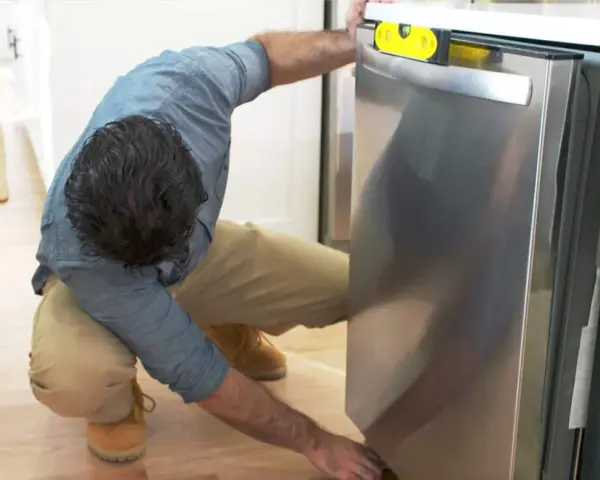 Man leveling a stainless steel dishwasher with a yellow bubble level, part of a guide on how to replace a dishwasher.