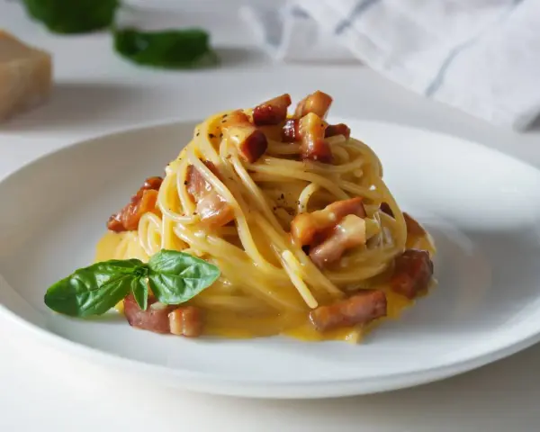 Pyszna carbonara z makaronem spaghetti, boczkiem i bazylią. W tle widać kawałek parmezanu, sugerując autentyczne carbonara skladniki.