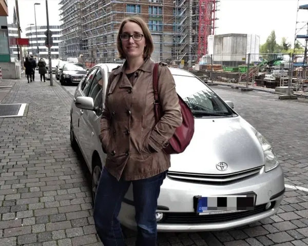 Frau mit brauner Lederjacke und roter Tasche steht neben silbernem Auto. Baustelle im Hintergrund. Sie ist free now fahrer.