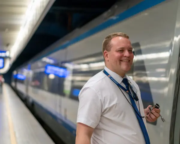 Uśmiechnięty konduktor PKP Intercity w białej koszuli i niebieskim krawacie stoi na peronie obok pociągu.