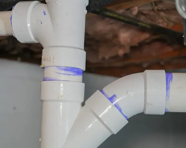 Close-up of various PVC pipe types, showing a vertical pipe connected to a horizontal pipe with a curved fitting, all marked with purple solvent cement.