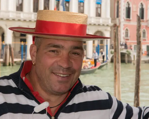 Un gondoliere sorridente, con il suo cappello iconico, pronto a navigare i canali di Venezia, la città che porta il soprannome di "La Serenissima".