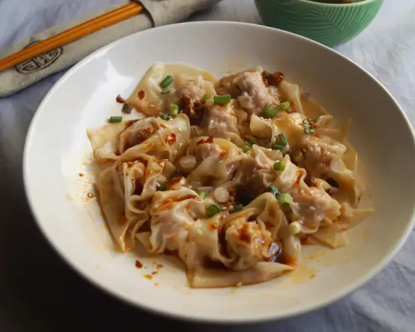Pyszne ciasto wonton polane pikantnym sosem, posypane szczypiorkiem i sezamem. Obok pałeczki i miseczka z sosem.