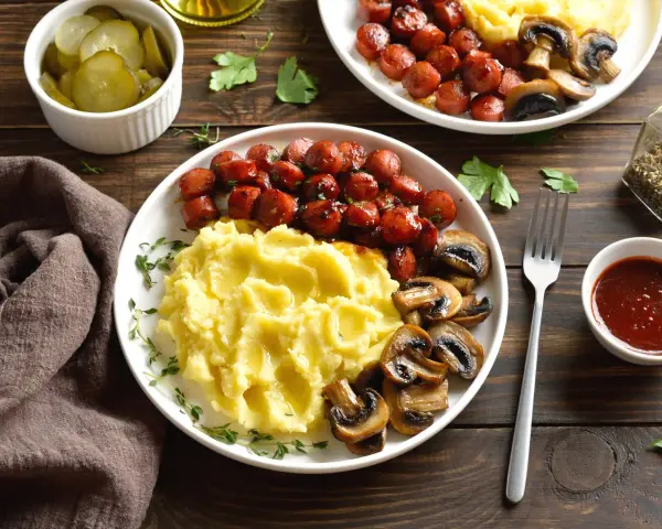 Talerz z purée ziemniaczanym, kiełbaskami i pieczarkami, obok miseczka z piklami i sosem.