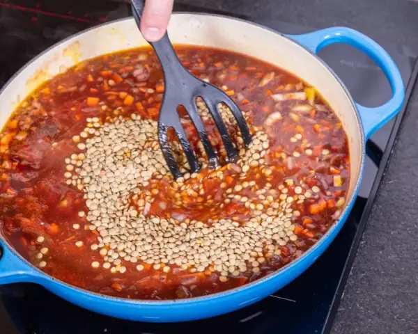 Eine Person rührt Linsen in eine köchelnde Bolognese-Sauce in einem blauen Topf.
