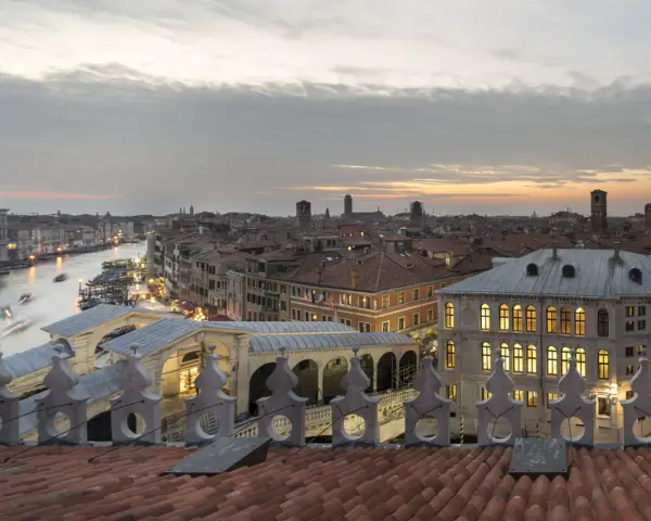 Vista dal **fondaco dei tedeschi Venezia terrazza** al tramonto, con il Canal Grande e il Ponte di Rialto illuminati.