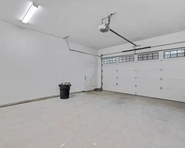 Nowy garaż z płyt betonowych projekt. Biała brama segmentowa, drzwi, czarny kosz na śmieci i sufitowe oświetlenie.