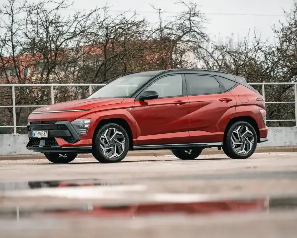 Czerwony Hyundai Kona Hybrid z czarnym dachem stoi na mokrym parkingu. Elegancki design i nowoczesne linie podkreślają jego hybrydowy charakter.