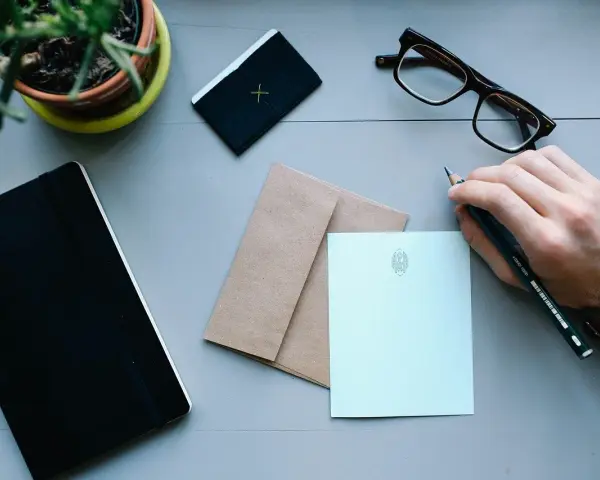 Dłoń z ołówkiem nad papierem, gotowa do napisania zwrotów do adresata. Obok notes, okulary i koperta.