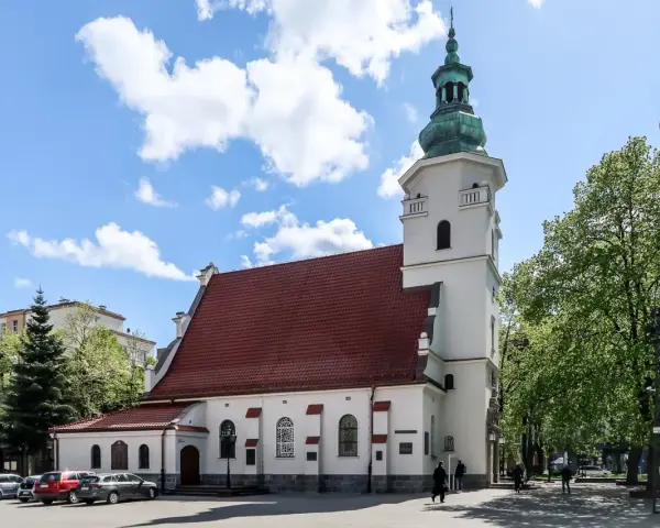 Biała bazylika pw. NMP Królowej Polski z czerwoną dachówką i zieloną wieżą, otoczona drzewami i budynkami.