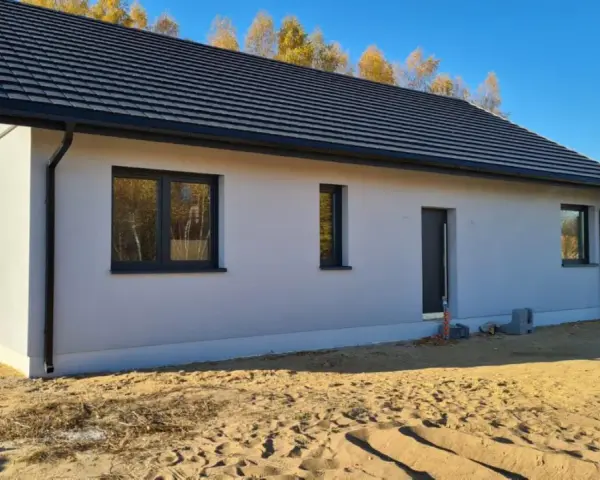 Nowy dom w stanie surowym zamkniętym, z szarym dachem i białymi ścianami, otoczony piaskiem i jesiennymi drzewami.