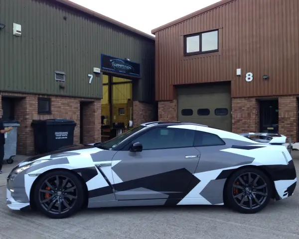 A Nissan GT-R with a striking geometric camouflage vinyl wrap for cars, parked outside a building with "WRAPTITION" signage.