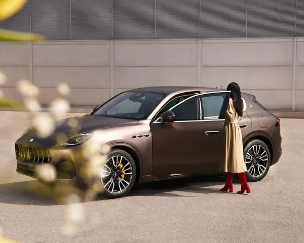 Elegancka kobieta w beżowej sukience i czerwonych kozakach zbliża się do brązowego Maserati SUV.