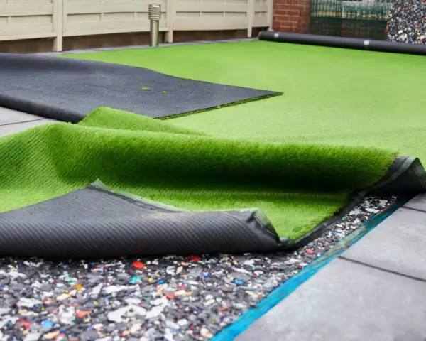 Instalación de césped artificial en terraza. Se ve el rollo de césped desplegado sobre una base de goma reciclada y baldosas.