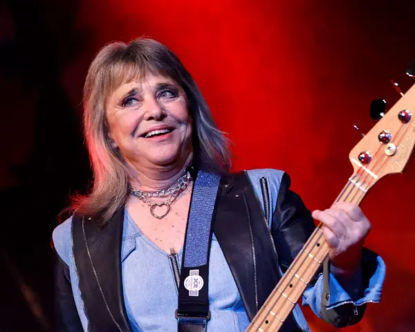 Suzi Quatro, die Rock-Ikone, spielt Bassgitarre. Sie trägt eine Lederjacke und eine Kette mit Herzanhänger. Ihr Alter ist nicht ersichtlich, aber ihre Energie ist spürbar.