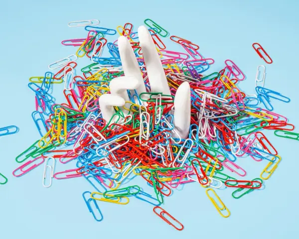 White hands emerge from a pile of colorful paperclips, symbolizing the struggle of how to stop being a workaholic and escape the daily grind.