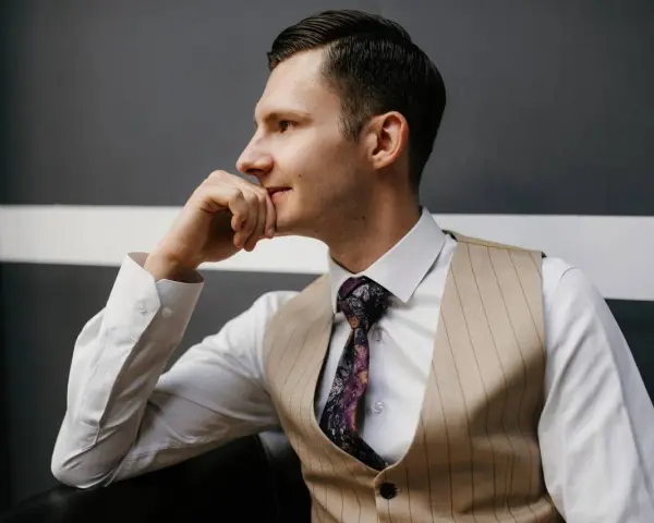 Elegancki mężczyzna w kamizelce, co zamiast marynarki, z dłonią na brodzie, wpatrzony w dal.