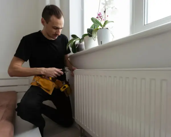 A man in a tool belt inspects a white radiator, checking for any signs of a cracked radiator before winter sets in.