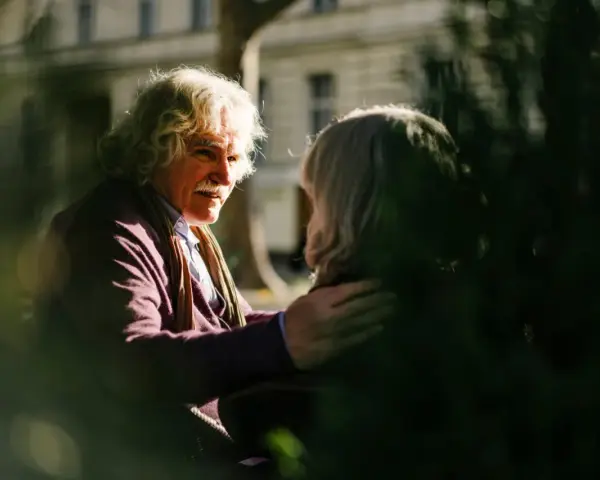 A man with white, curly hair and a mustache gently places his hand on another person's shoulder, offering a comforting response. This scene illustrates react vs respond examples.