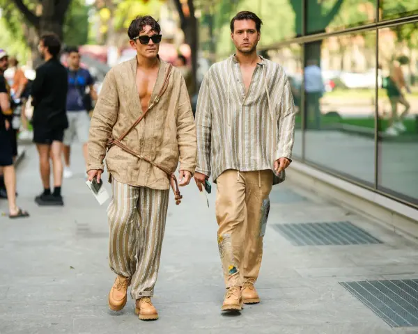 Dos hombres pasean con estilo. Uno lleva un kimono beige y pantalones de lino a rayas. El otro, una camisa a rayas y pantalones beige. Un ejemplo de cómo combinar pantalón lino beige hombre.