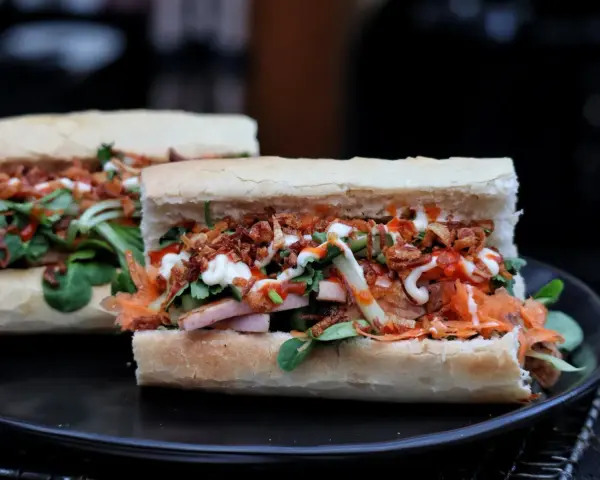 Zwei appetitliche Banh Mi Sandwiches mit Fleisch, Gemüse, knusprigen Zwiebeln und Saucen auf einem Teller.