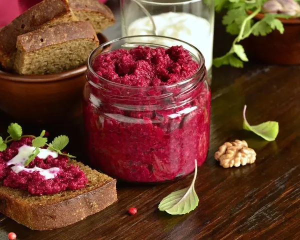 Glas mit leuchtend rotem Rote Bete Pesto, daneben Brot mit Pesto und Joghurt. Walnuss und Kräuter garnieren die Szene.