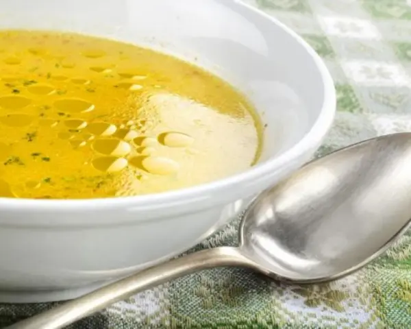 Una ciotola bianca piena di brodo di carne dorato, con goccioline d'olio in superficie. Un cucchiaio d'argento è appoggiato accanto.