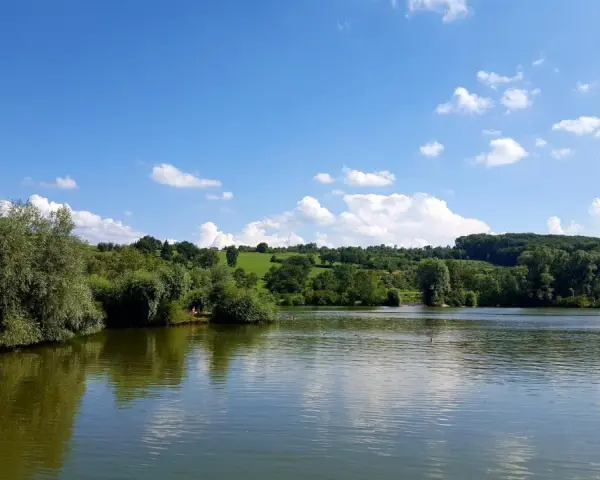 Ein sonniger Tag am Badesee Esslingen. Grüne Hügel, Bäume und ein blauer Himmel mit weißen Wolken spiegeln sich im ruhigen Wasser.