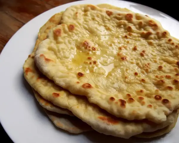 Stos świeżo upieczonych, lekko przypieczonych placków roti i naan, polanych roztopionym masłem, na białym talerzu.