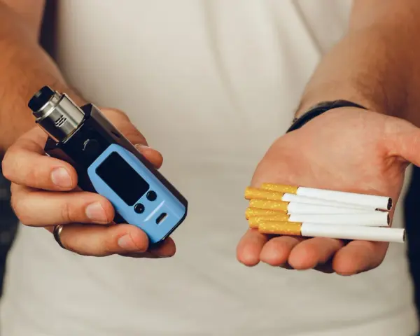 Man holds a vape in one hand and cigarettes in the other, posing the question: que es mejor el vaper o el tabaco.