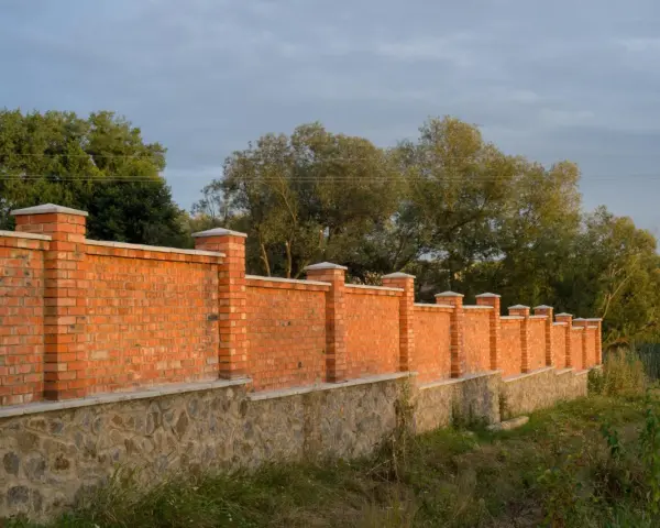 Solidny płot murowany z cegły i kamienia, z ozdobnymi słupkami, otacza teren porośnięty zielenią.
