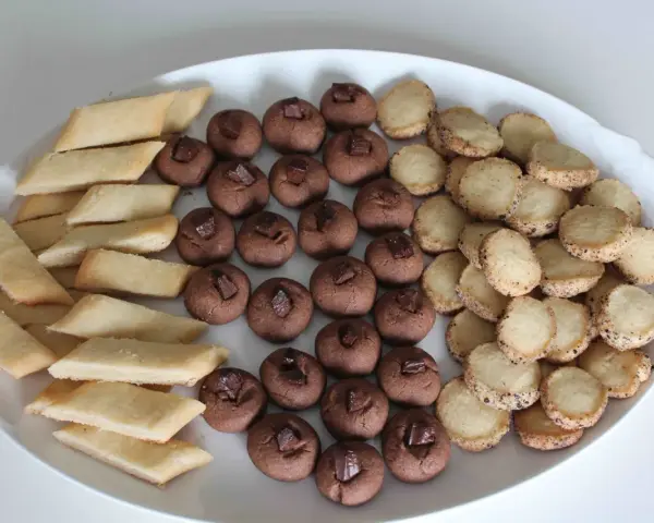 En vit oval bricka fylld med tre sorters hembakade småkakor: rombiska, chokladpraliner och runda med strössel. Perfekt för fika!