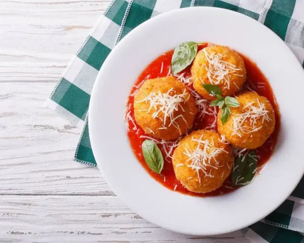 Quattro arancini di riso dorati, cosparsi di parmigiano e serviti su un letto di salsa di pomodoro, guarniti con basilico fresco.