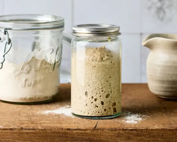Blubbernder Sauerteig im Glas, bereit zum Backen. Daneben ein Glas mit Mehl, das zeigt, wie man den Sauerteig pflegen kann.