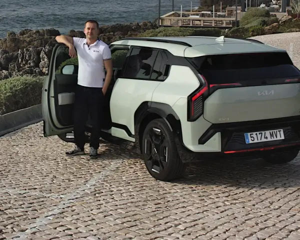 Mężczyzna opiera się o drzwi nowego, jasnoszarego Kia EV3, zaparkowanego na brukowanej drodze nad morzem.