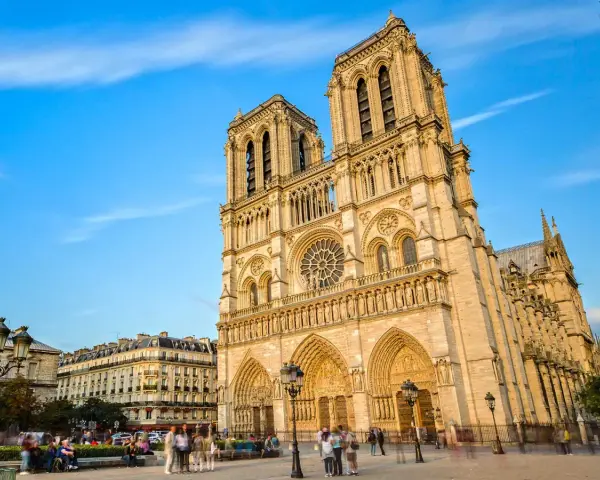 La majestueuse façade de Notre Dame de Paris se dresse sous un ciel bleu, ornée de détails gothiques. Des passants flânent sur la place.
