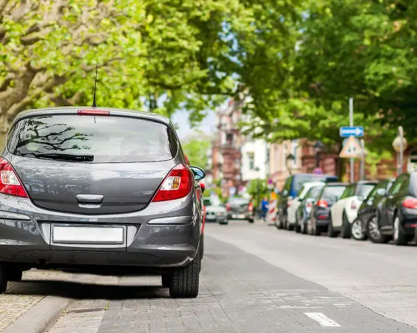 Graues Auto parkt am abgesenkten Bordstein. Mehrere Autos reihen sich die Straße entlang unter grünen Bäumen.
