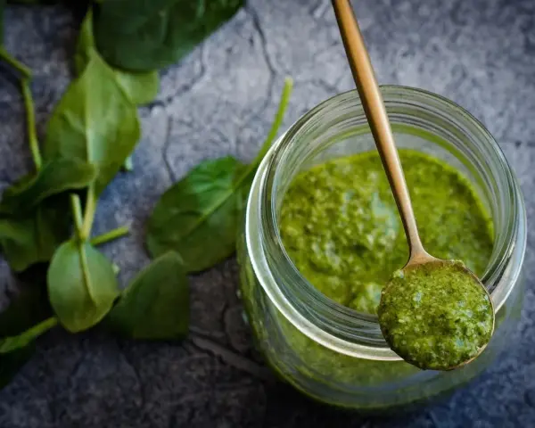 Słoik z zielonym pesto, na łyżce porcja sosu. Czy pesto ze słoika się podgrzewa? Świeże liście bazylii obok.