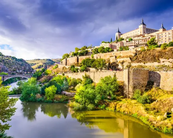 El **río Tajo** serpentea al pie de Toledo, una ciudad amurallada coronada por un imponente alcázar. Un puente de piedra cruza el agua, conectando las verdes orillas.