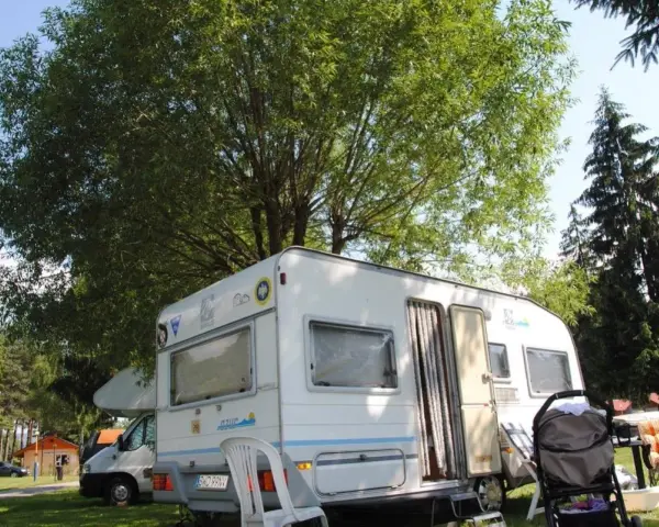 Przyczepa kempingowa na polu namiotowym Jonidło, otoczona zielenią. Obok wózek i leżak.