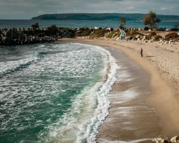 Piękne, turkusowe morze w Bułgarii obmywa piaszczystą plażę. W oddali widać wzgórza i samochody.