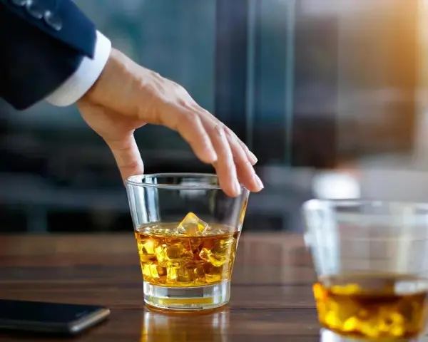 Hand greift nach einem Glas goldbraunen Whiskey mit Eiswürfeln. Ein Genuss, auch für Menschen, die glutenfreien Whiskey bevorzugen.